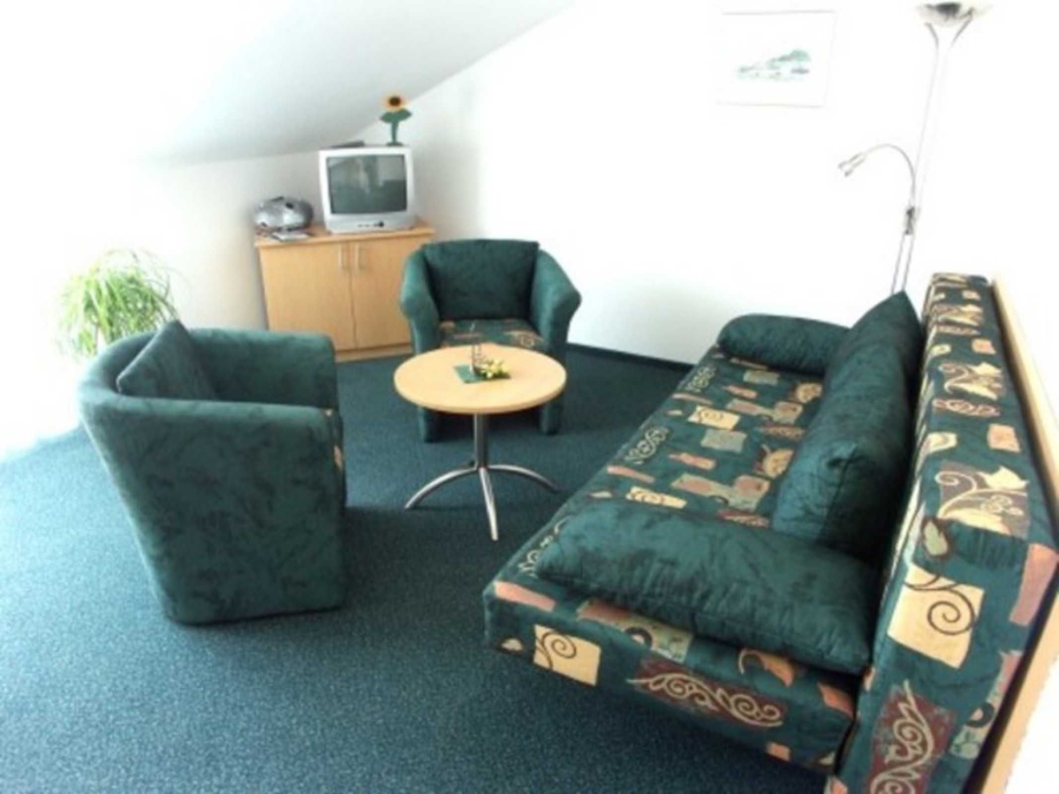 A small, cozy room with a green sofa, two armchairs, a small table, and a television on a cabinet.