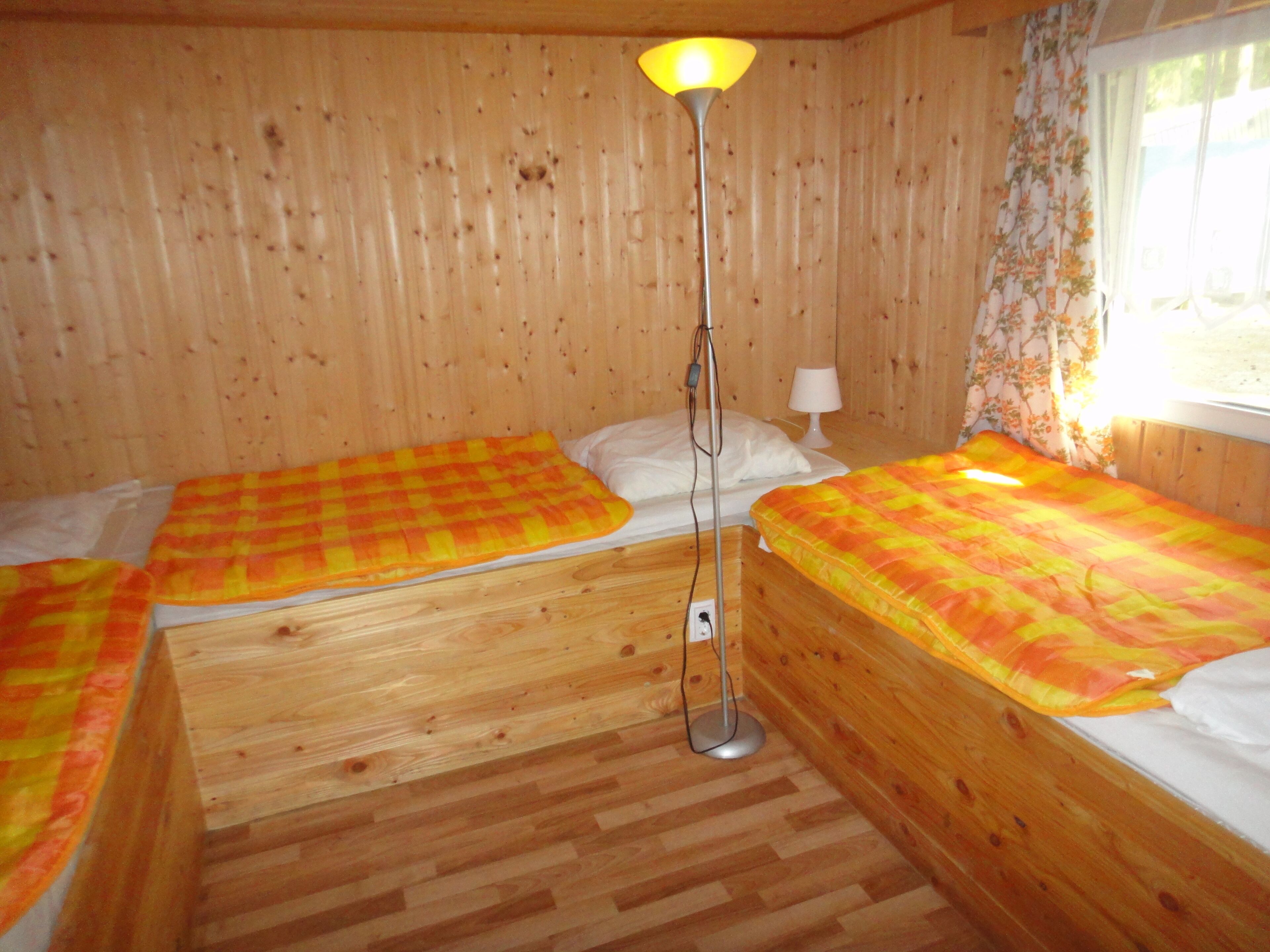 Ein Holzraum mit zwei Betten, jedes mit einer karierten Decke, einer Stehlampe und einem Fenster mit blumigen Vorhängen.