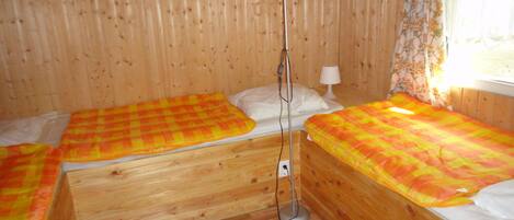 Ein Holzraum mit zwei Betten, jedes mit einer karierten Decke, einer Stehlampe und einem Fenster mit blumigen Vorhängen.