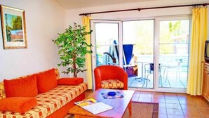 A living room with a sofa, a table, a chair, a plant, and a view through a glass door.