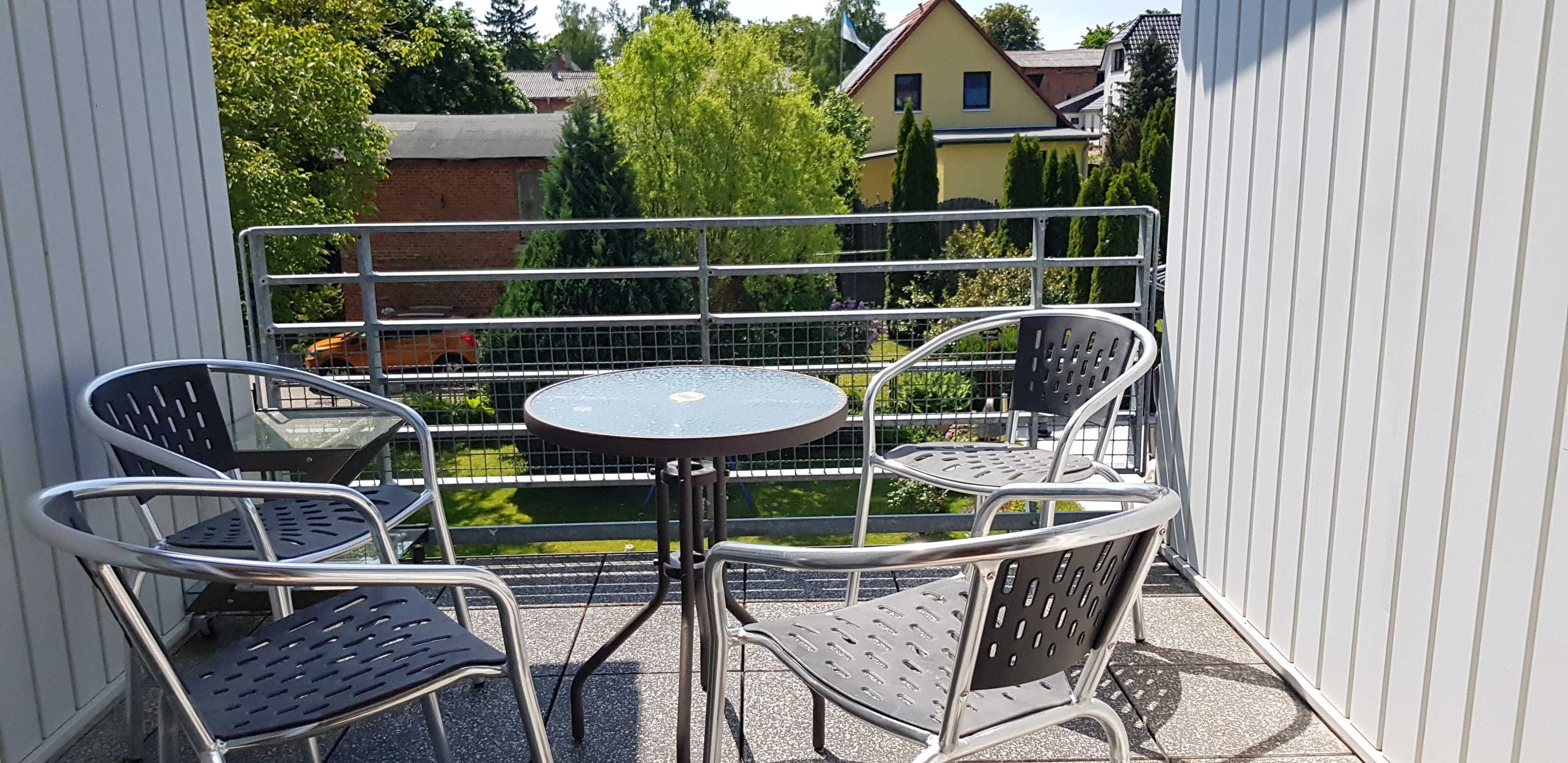 En balkon med et rundt bord og fire stole, gelænder i metal og udsigt til huse og træer.