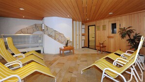 Ein Zimmer mit Holzwänden, einer Steinwand und einer Sauna. Gelb gestreifte Liegestühle und ein kleiner Tisch mit einer Vase sind vorhanden.
