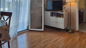 Quarto com piso de madeira, uma TV em um armário branco, uma luminária e uma porta de vidro deslizante que dá para uma varanda.