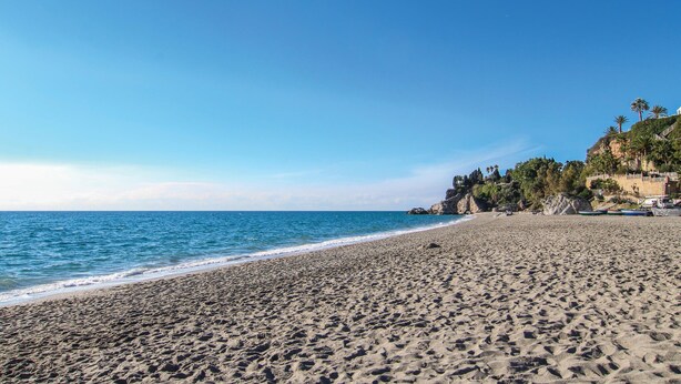 Ein weiter Sandstrand mit sanften Wellen, einem klaren blauen Himmel und einem felsigen, bewachsenen Felsen am rechten Rand.
