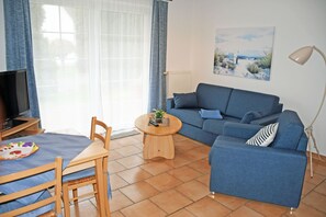 Uma sala de estar com um sofá azul, uma mesa de centro redonda de madeira, uma televisão em um móvel e um quadro na parede.