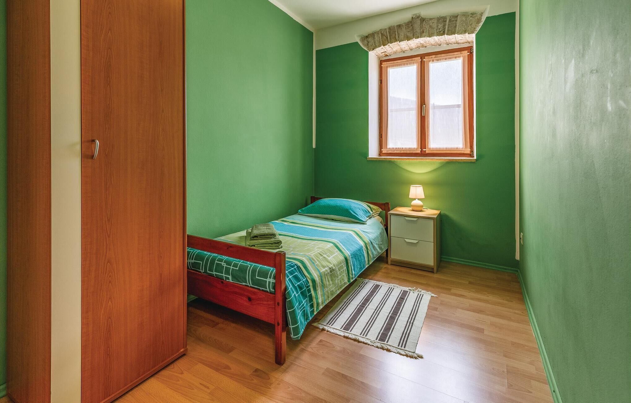 A small, single-bed room with green walls, a wooden door, a window with a curtain, a bedside table with a lamp, and a striped rug.