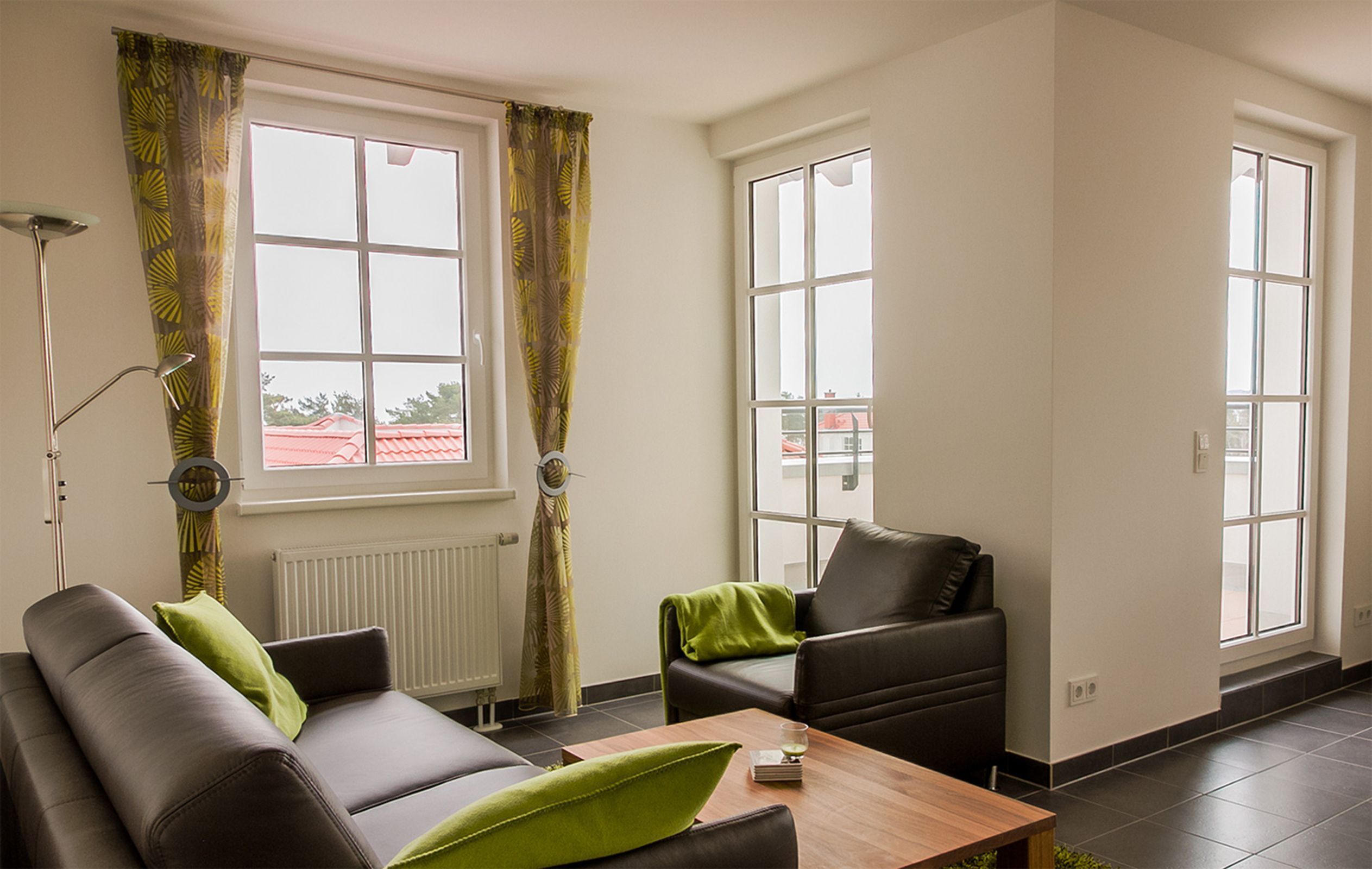 A modern living room with a sofa, a coffee table, and a chair. There are large windows with curtains, a floor lamp, and a radiator.