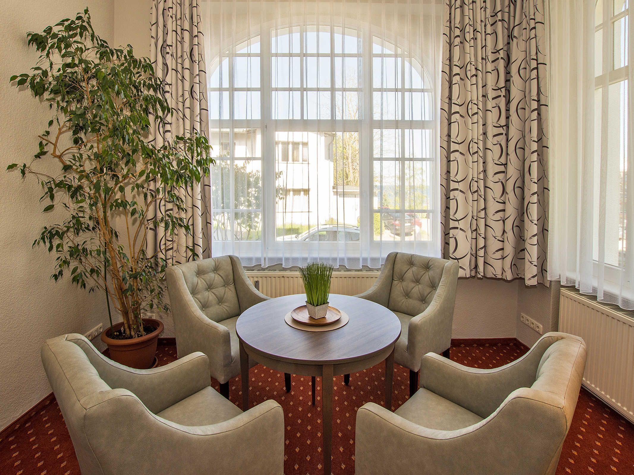 A room with a round table, four armchairs, a potted plant, and a large window with curtains.