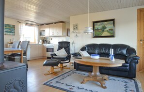 A living room with a black leather sofa, a wooden coffee table, a dining area with wicker chairs, a kitchen with wooden cabinets, and a painting on the wall. - 3 bedroom beach front home in Hjørring (Hjørring)