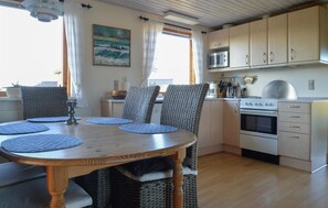 A dining area with a round wooden table, wicker chairs, and a kitchen with white cabinets and appliances. - 3 bedroom beach front home in Hjørring (Hjørring)