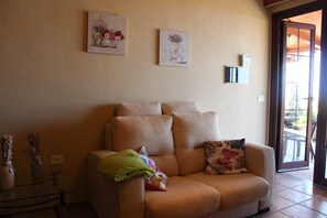 A living room with a beige sofa, two wall-mounted artworks, a green throw blanket, and a glass coffee table with vases. - Casa Cueva de las Palomas (Los Llanos)