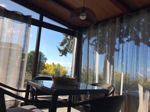 Una stanza luminosa con un tavolo in vetro e sedie in vimini, ampie finestre con tende leggere e un lampadario di vimini.