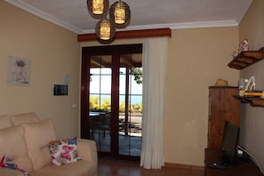 A living room with a beige sofa, a television on a wooden stand, and a view through a glass door to an outdoor area.