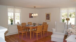 A dining room with a wooden table set for six, surrounded by wicker chairs. There is a white armchair and a small white table with a lamp.
