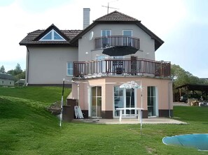 Una casa de dos pisos con un balcón, una estructura adosada pequeña con una mesa y sillas, y una piscina al fondo.