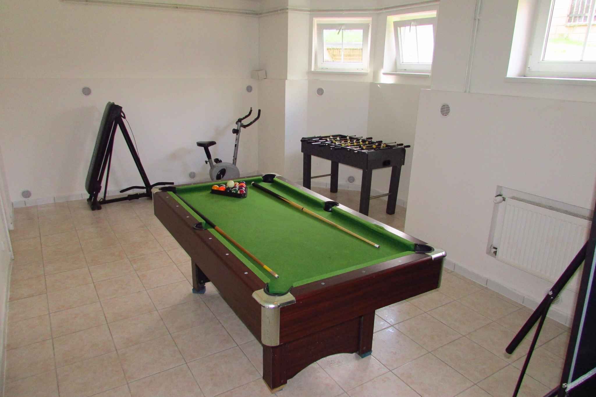 A pool table with a green felt surface, a foosball table, and a stationary exercise bike in a room with white walls and tiled flooring.