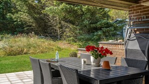 An outdoor patio with a table and chairs, a grill, and a garden in the background.
