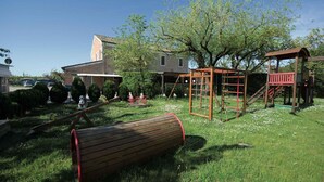 Ein Spielplatz mit Schaukeln, einer Rutsche und einer Kletterkonstruktion, umgeben von Rasen, Bäumen und einem Haus im Hintergrund.