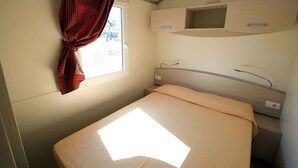 Ein kleines, gut beleuchtetes Schlafzimmer mit einem Einzelbett, einem Fenster mit Vorhängen und einem fest eingebauten Regal über dem Bett.