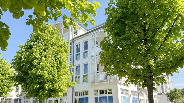 A white building with large windows and balconies, surrounded by lush green trees.