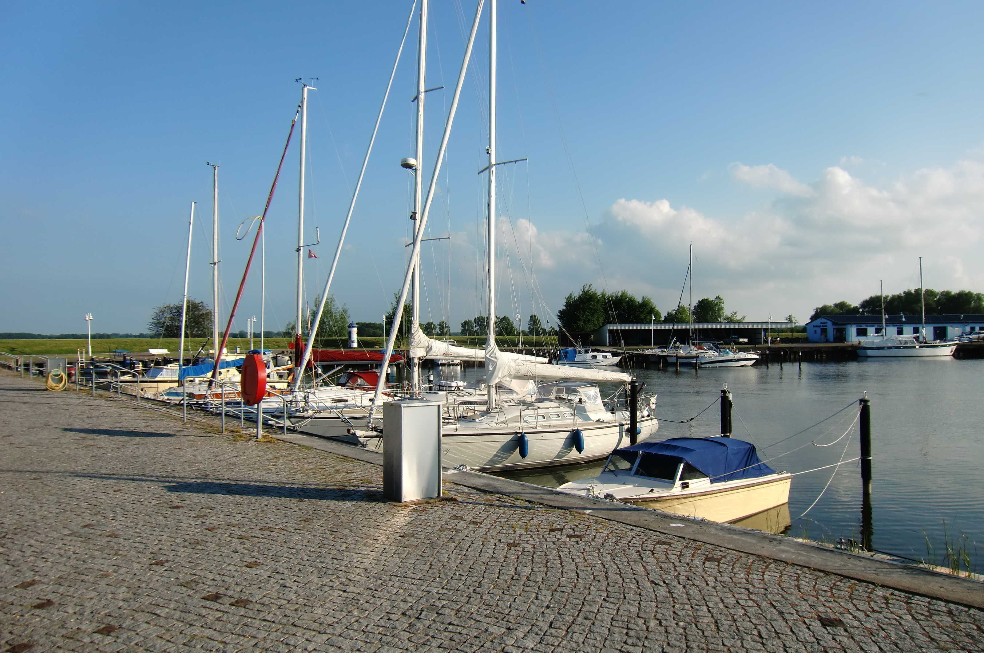Un port de plaisance où plusieurs voiliers sont amarrés le long d’une promenade au bord de l’eau pavée de pierres.
