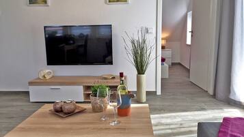 Ein modernes Wohnzimmer mit Flachbildfernseher, Couchtisch aus Holz, Zimmerpflanzen und einer gemütlichen Sitzecke.