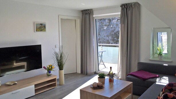 A modern living room with a flat-screen TV, a wooden coffee table, a grey sofa, and a balcony with a table and chairs. - Le Papillon apartment rental - Apartment (Kröslin)