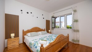 Een slaapkamer met een houten bed, een nachtkastje met een lamp, een raam met gordijnen en wanddecoraties, waaronder bloemen en een vlinder.