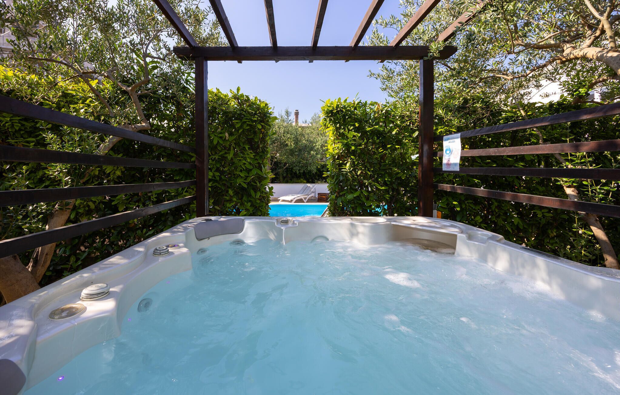 Ein Whirlpool, umgeben von einer Holzpergola und üppiger Vegetation, mit einem Pool im Hintergrund.