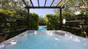 Een hottub omringd door een houten pergola en weelderige begroeiing, met een zwembad op de achtergrond.