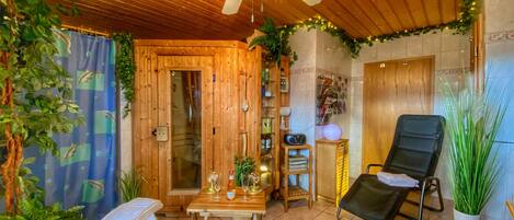 Un intérieur en bois avec un ventilateur de plafond, une porte en bois, une petite table avec des verres à vin, un fauteuil noir et des plantes en pot.
