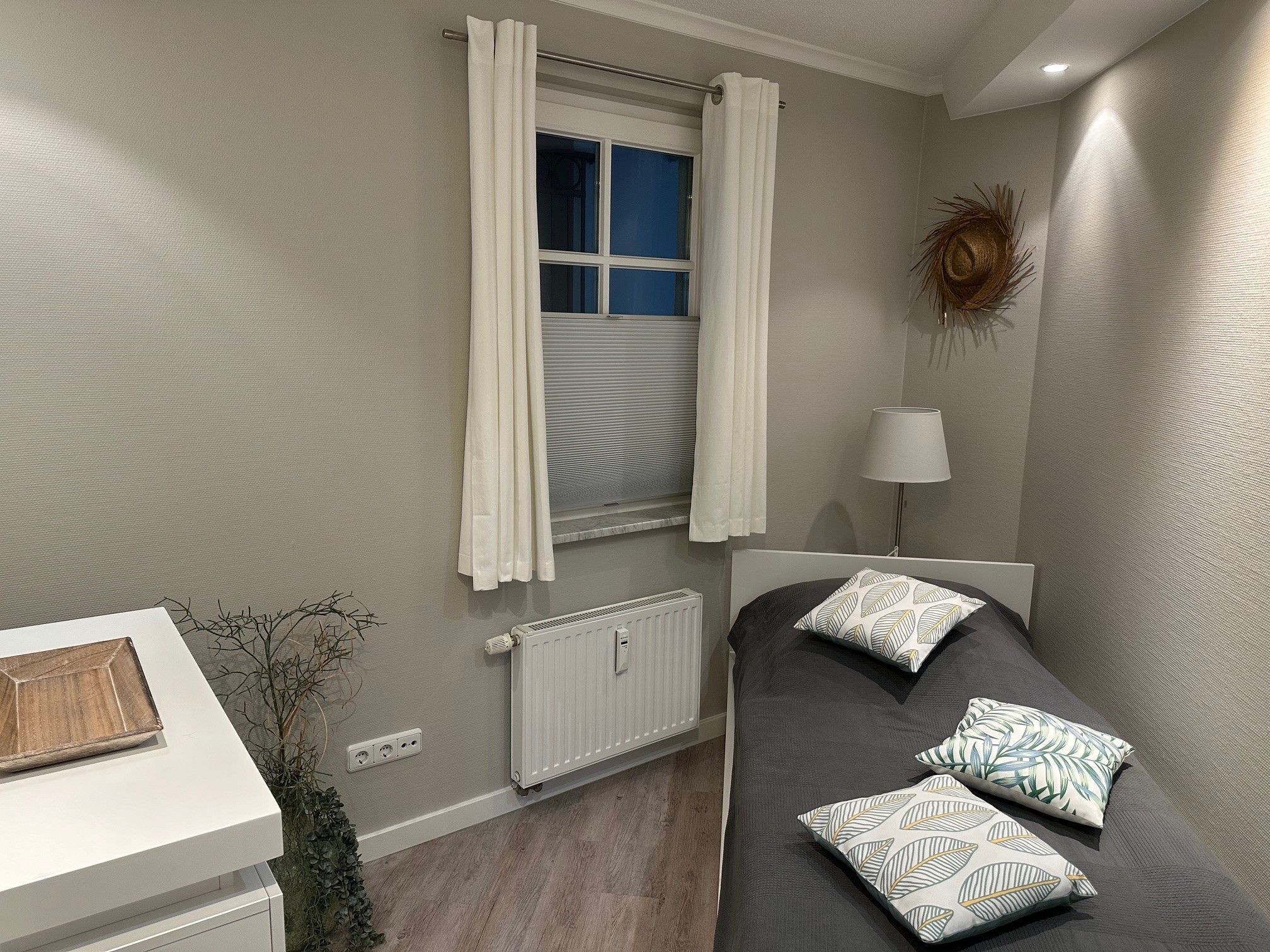A bedroom with a bed, a window with curtains, a radiator, a bedside lamp, and a wall decoration.