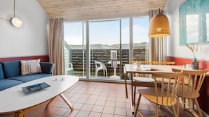 Ett modernt rum med en soffa, ett matbord med stolar, en hängande lampa och ett stort fönster med utsikt över omgivningen.