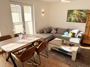 A modern living room with a wooden dining table, brown chairs, a grey sectional sofa with colorful cushions, a coffee table, and a painting of a forest.