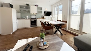 A modern kitchen with white cabinets, a microwave, and a TV above the counter. There is a dining table with chairs and a small potted plant on the table.