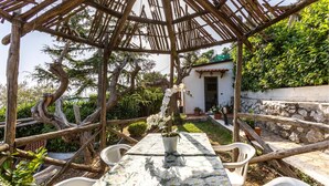 Una estructura de pérgola de madera con una mesa y sillas debajo, rodeada de abundante follaje y un muro de piedra.