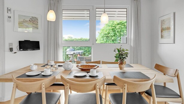 A dining area with a table set for four, surrounded by wooden chairs, a television mounted on the wall, and large windows with curtains.