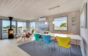 A modern dining area with a large table, colorful chairs, a wood-burning stove, and large windows offering views of the outdoors. - Lovely home in Fanø with WiFi (Fanø)