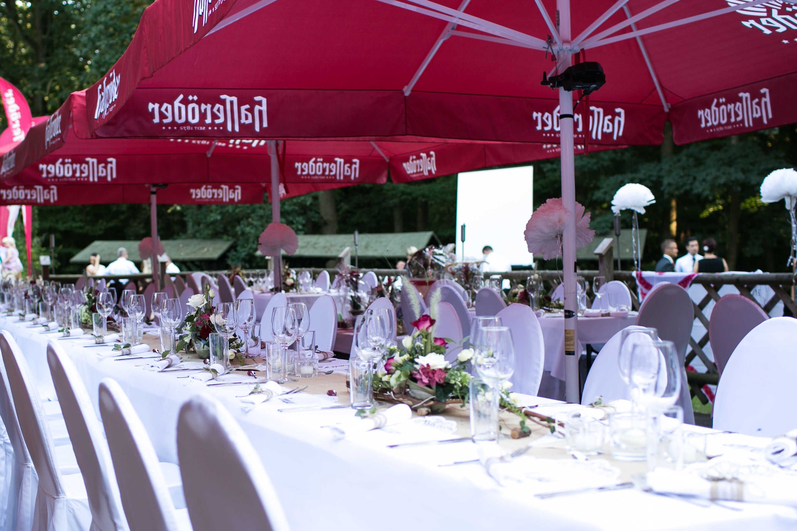 A banquet table set with white tablecloths, red umbrellas, and white chairs.