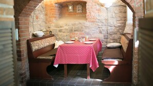 A stone-walled dining area with a brick arch, wooden benches, a table with a red tablecloth, and a stone fireplace.