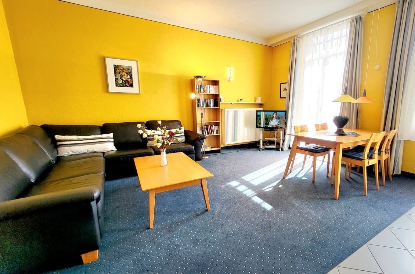 A living room with a black sofa, a wooden coffee table, a dining table with chairs, a bookshelf, and a television.