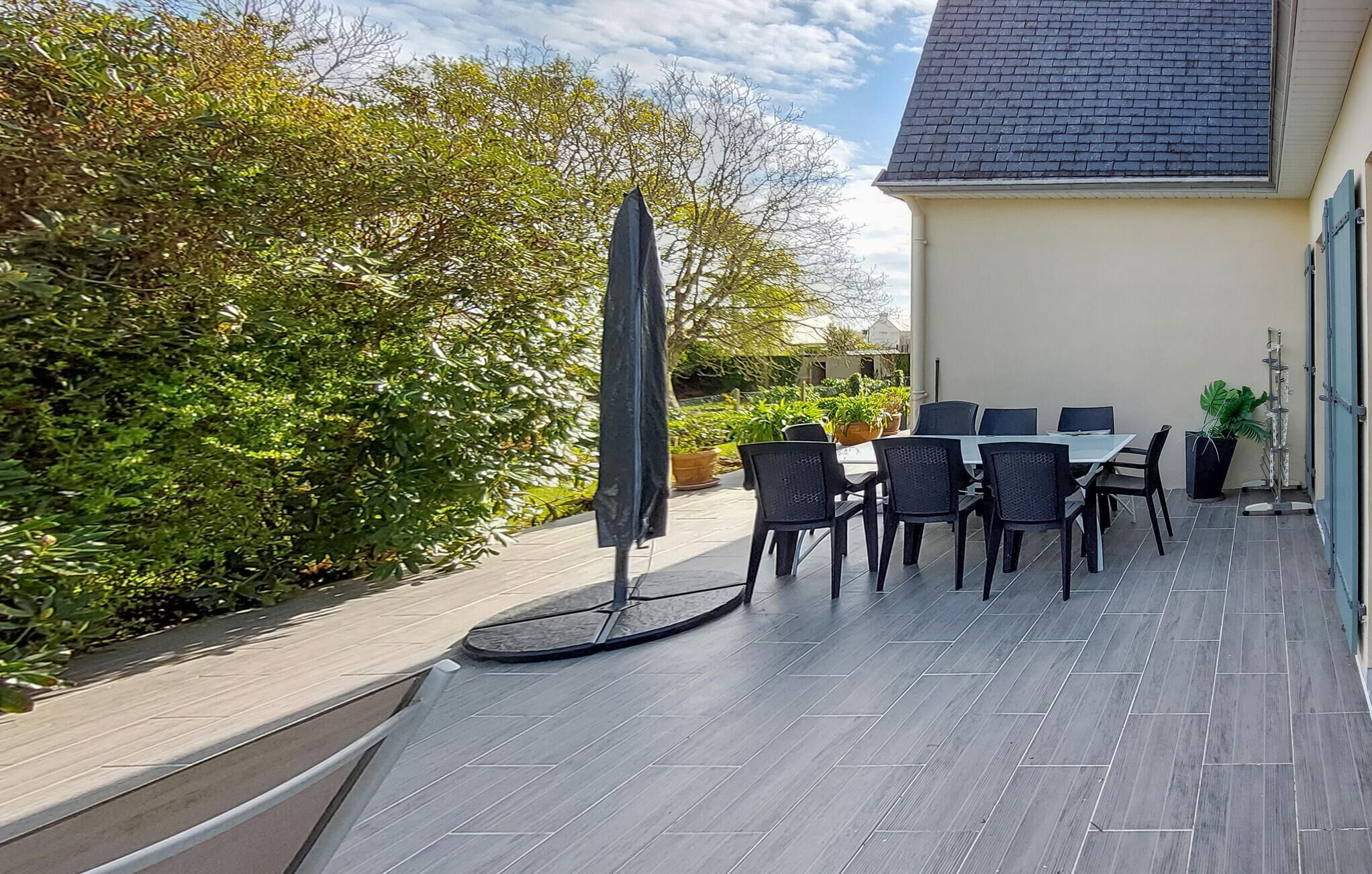 Une terrasse extérieure avec une table et des chaises, entourée de verdure et bordée d’un bâtiment dont le toit est recouvert de tuiles.