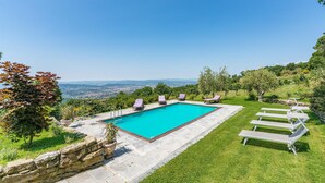 A rectangular pool with a clear blue water, surrounded by a stone wall and grass, with lounge chairs and a view of distant hills and a city.