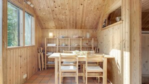 A wooden interior with a dining table and chairs, shelves with dishes, and a window with curtains.