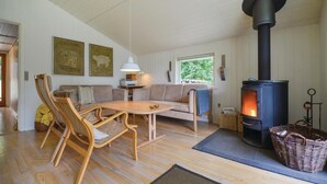 A cozy living room with a wooden table, two sofas, a large black wood-burning stove, and a window with a view of greenery. - Lovely home in Ebeltoft with sauna (Ebeltoft)