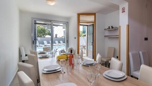 Een eetgedeelte met een houten tafel gedekt voor vier personen, omringd door witte stoelen. Er is uitzicht op een zwembad en ligbedden buiten.