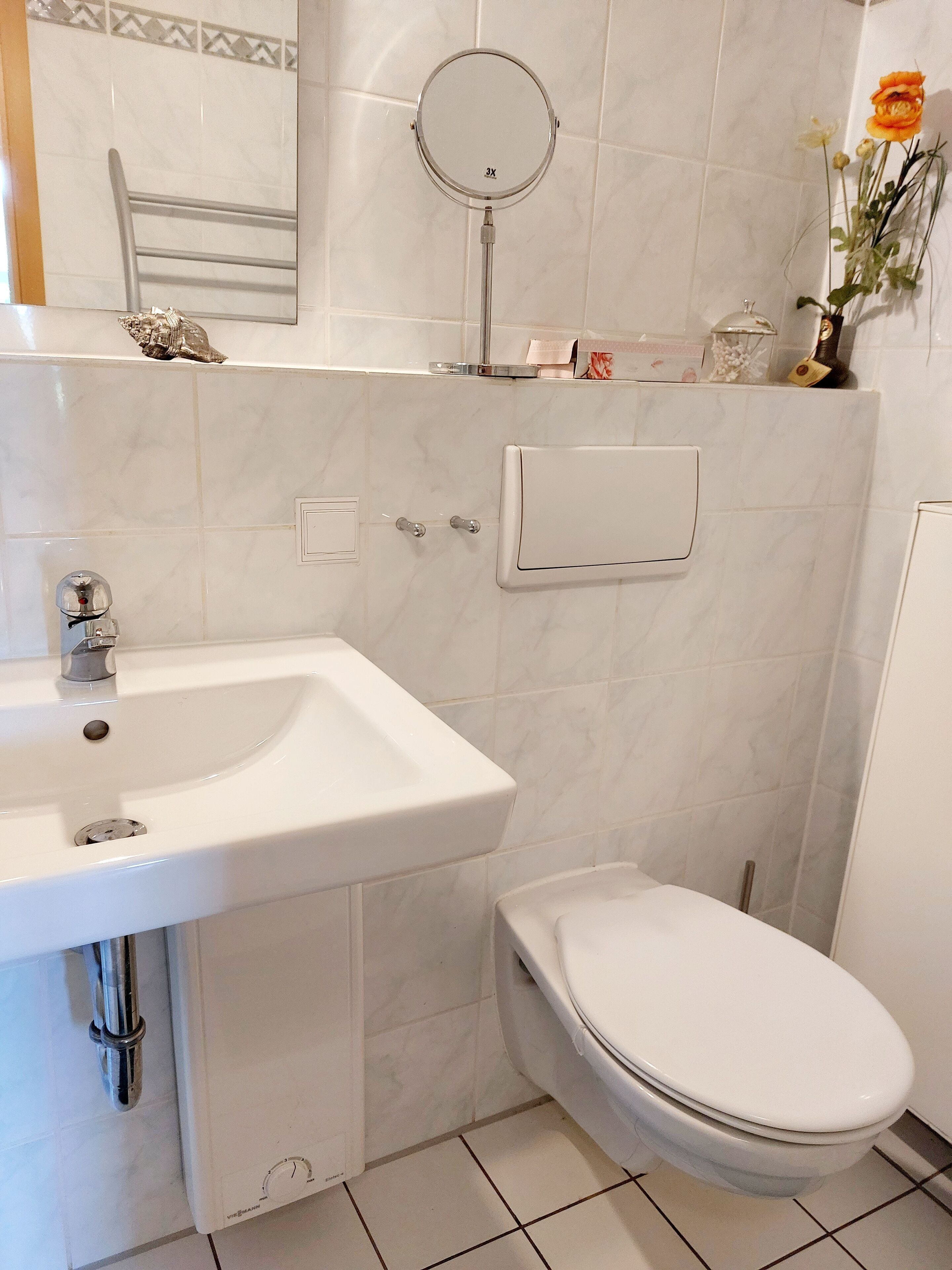 A bathroom with a white sink, toilet, and mirror. There is a shelf with toiletries and a vase with flowers.