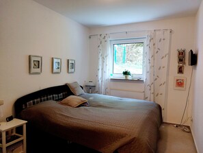 Quarto com cama, mesa de cabeceira, abajur, janela com cortinas e quadros na parede.