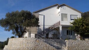 Une maison à deux étages, avec un escalier en pierre menant à l’entrée, entourée d’arbres et sous un ciel dégagé.