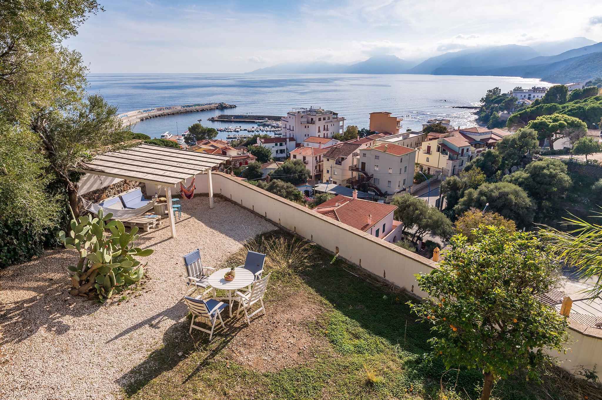A terrace with outdoor seating, a view of the sea, and buildings in the background.
